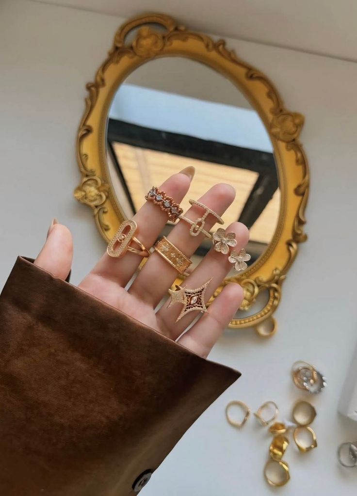 Hand adorned with gold rings, reflected in a vintage ornate mirror. Elegant jewelry display showcasing various ring designs.