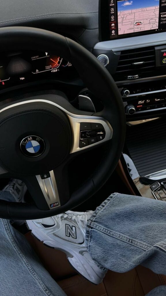 Inside view of BMW car dashboard with steering wheel, displaying navigation system; foot in white sneaker resting casually on the seat.