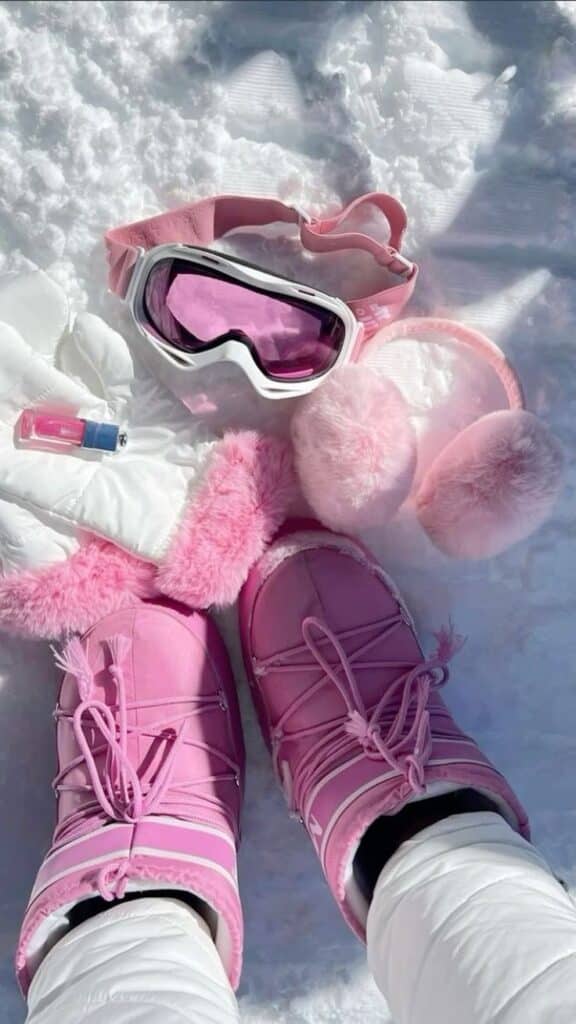 Pink winter accessories on snow: boots, ski goggles, earmuffs, lipstick, and gloves. Cozy and stylish winter gear.