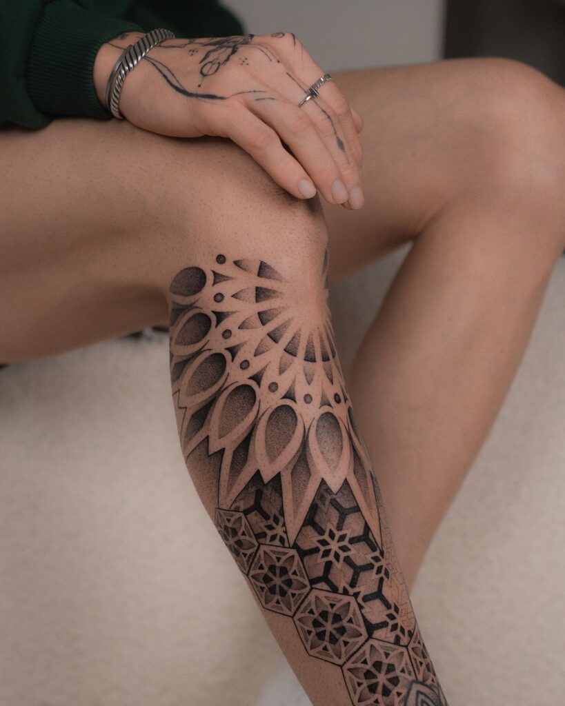 Intricate geometric tattoo design on leg and hand, featuring patterns and shading, with hand resting gently on knee.
