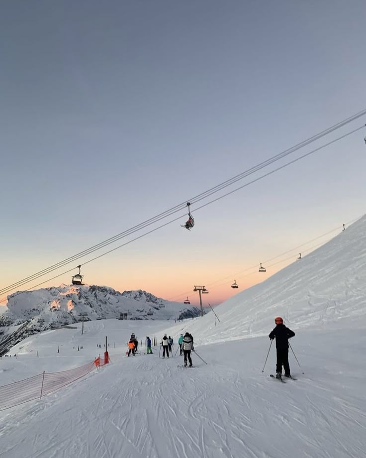 Skiers on snowy mountain slope at sunset with ski lifts overhead and panoramic view of snow-covered peaks and clear sky.