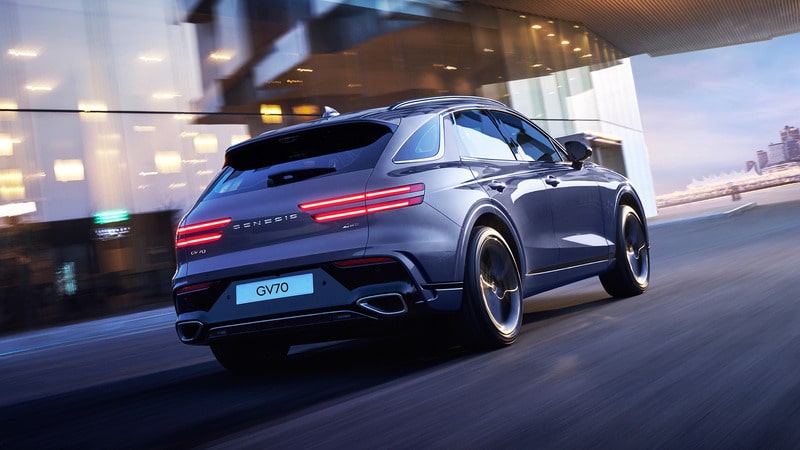 Sleek Genesis GV70 SUV driving swiftly in an urban setting with blurred city lights, showcasing modern design and performance.