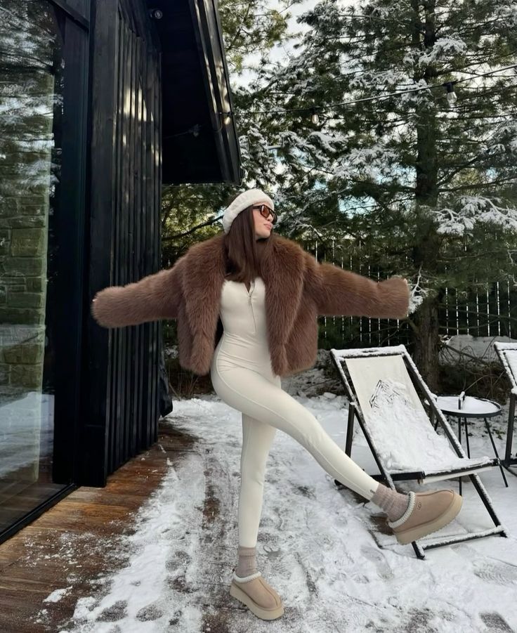 Woman in a fur coat and winter outfit posing outdoors in snowy setting, near a modern cabin and snow-covered trees.