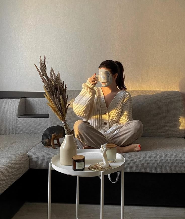 Cozy living room scene with person in sweater sipping coffee, sitting cross-legged on sofa, table with candles and dried flowers in foreground.
