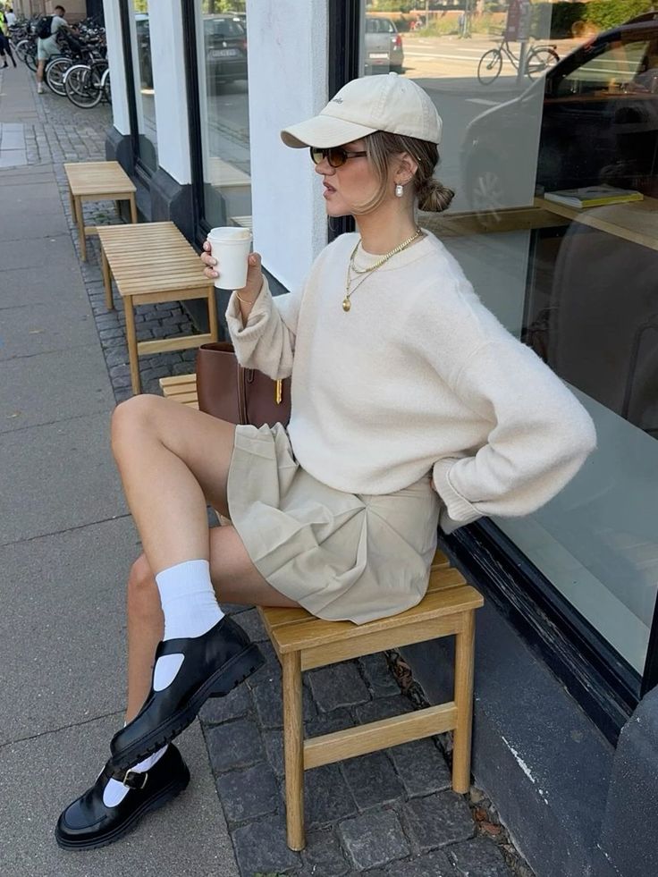 Fashionable person sitting outdoors, wearing a beige outfit, cap, and sunglasses while sipping coffee; street style inspiration.