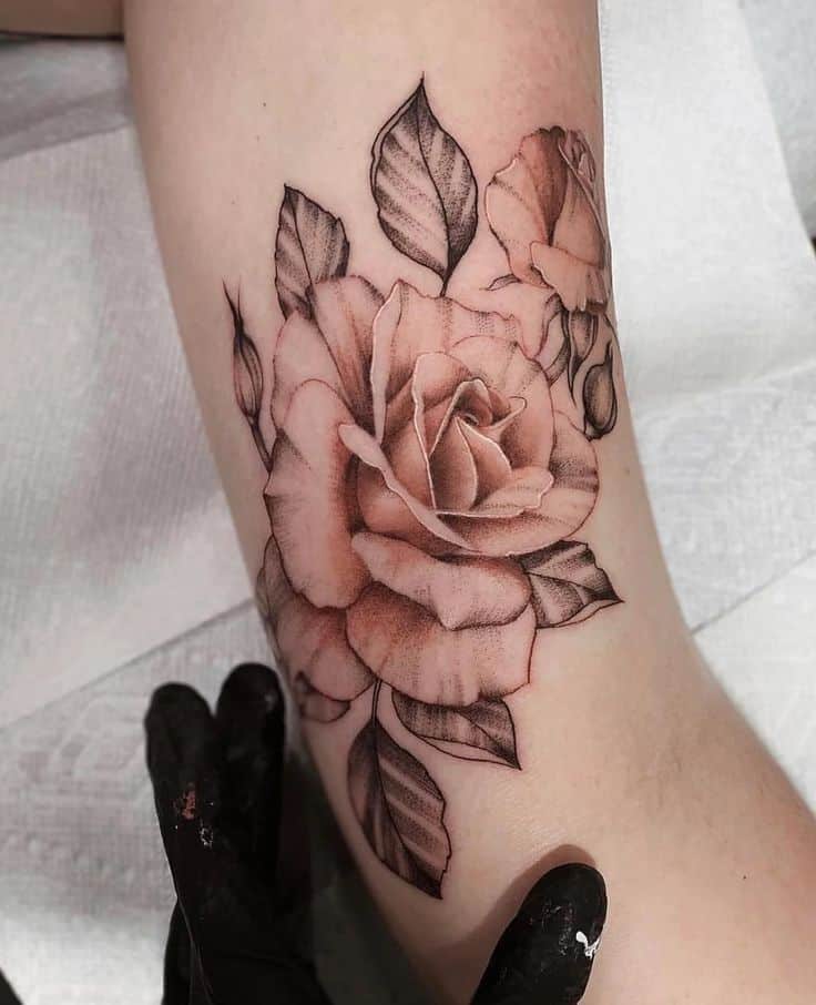 Realistic rose tattoo on arm, featuring detailed petals and shaded leaves, crafted by an artist wearing black gloves.