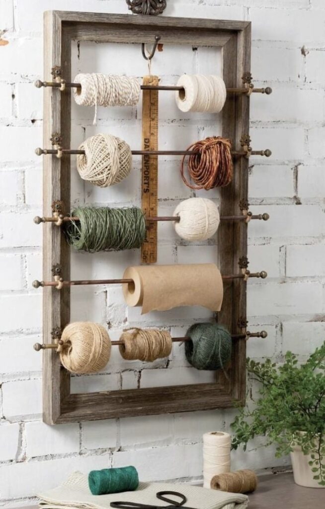 DIY twine organizer mounted on brick wall with wooden frame, holding various colored spools, a ruler, and a paper roll. Ideal for crafts.