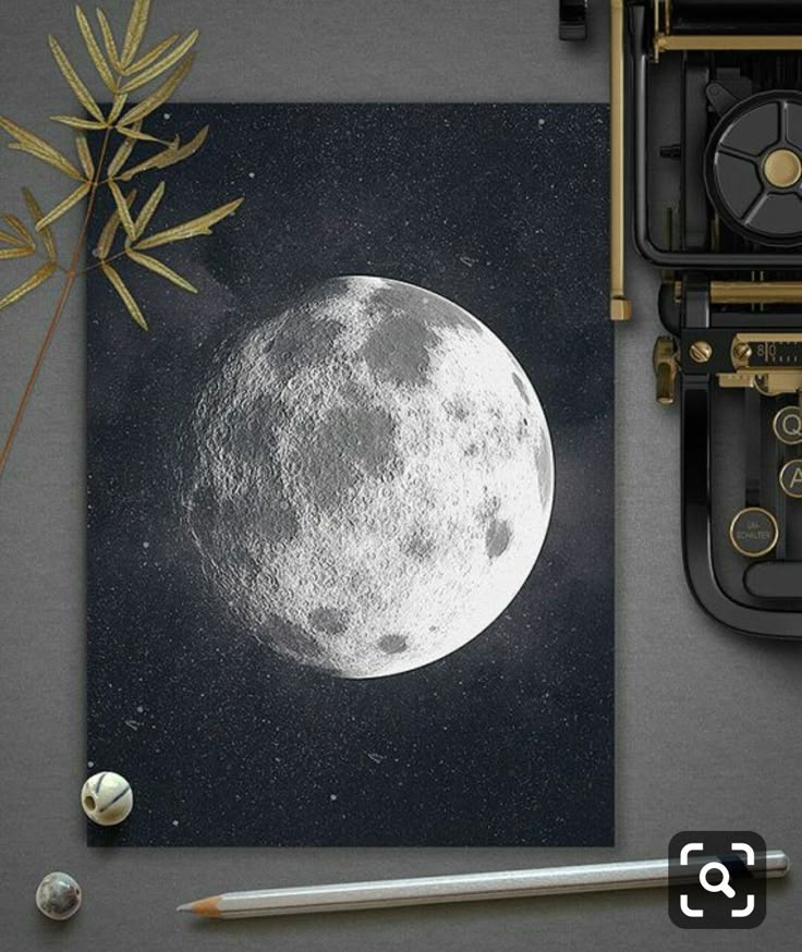 Print of the moon on a gray surface next to a pencil, dry leaf, and vintage typewriter; artistic celestial composition.