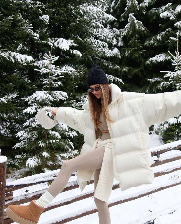 Woman in snowy forest wearing white winter coat, beige outfit, and boots, balancing playfully on a wooden fence.