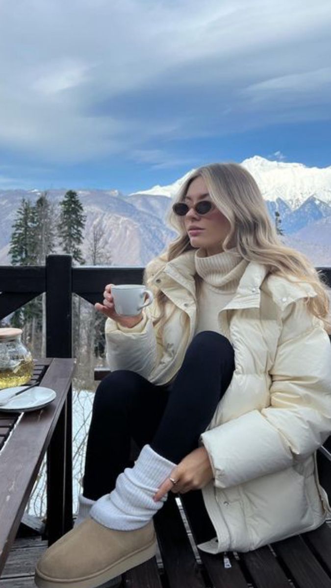 Woman in winter coat enjoying coffee on a mountain deck with snowy peaks in the background.