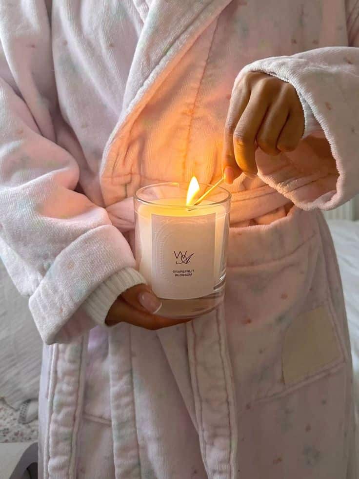 Person in cozy robe lighting a grapefruit blossom scented candle with a match, creating a warm, relaxing atmosphere.