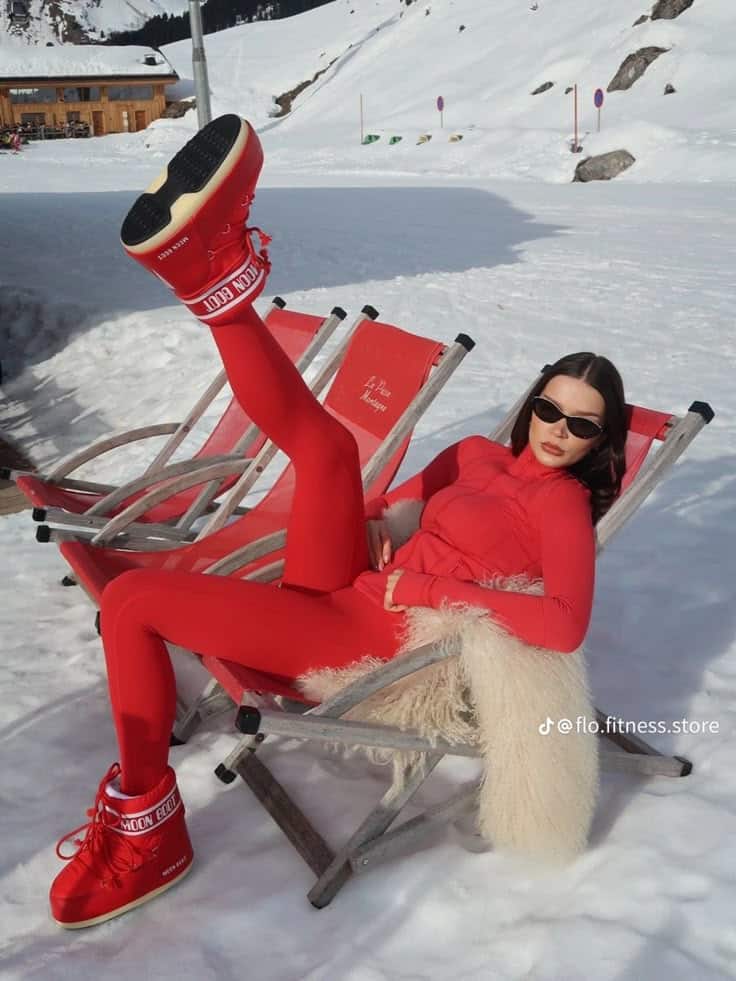 Person lounging in the snow wearing a red outfit and moon boots, striking a stylish pose on a deck chair.