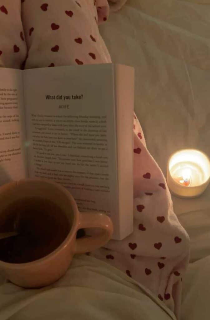 Cozy setup with an open book, tea mug, and candle, all on red heart-patterned pajamas for a perfect reading night.
