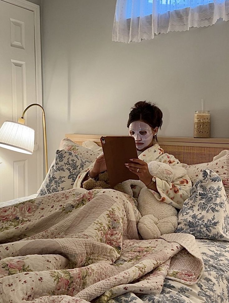 Person relaxing in bed with a face mask and tablet, surrounded by floral bedding and cozy decor, creating a serene nighttime routine.