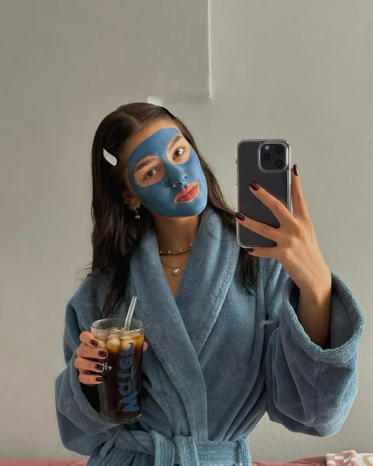 Woman in blue robe with face mask takes mirror selfie while holding iced drink, showcasing skincare routine and relaxation.