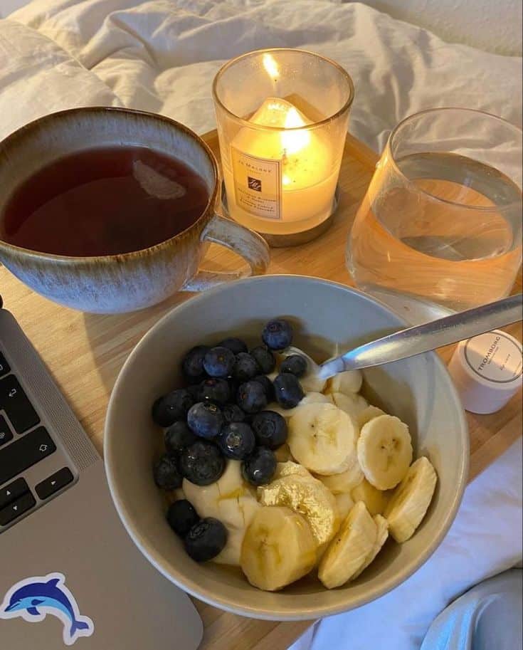 Cozy breakfast in bed with yogurt, blueberries, bananas, tea, candle, and laptop. Perfect for a relaxing morning routine.