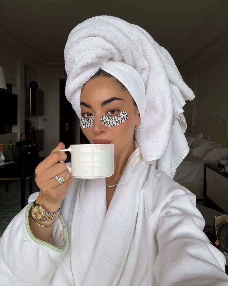 Woman in a robe and towel enjoying a cup of coffee with eye patches, relaxing morning routine in a cozy room.