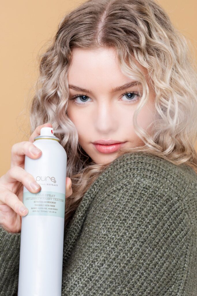 Woman with curly hair holding a spray can against a neutral background, showcasing hair product.