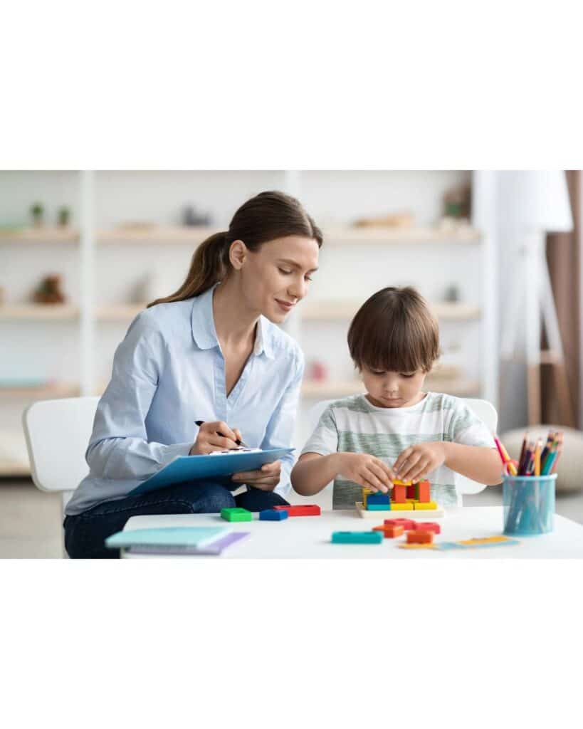 Child building with blocks while a woman observes and takes notes, highlighting learning and creativity in a classroom setting.