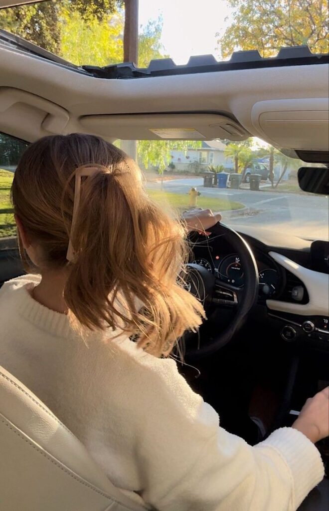 Person with ponytail driving a car, enjoying a sunny day, through an open sunroof; vibrant neighborhood in the background.