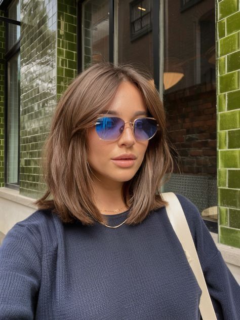 Woman with sunglasses and brown hair posing in front of green brick wall, wearing a navy blue sweater.