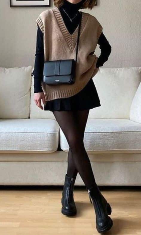 Fashionable outfit with a beige vest, black turtleneck, mini skirt, tights, and chunky boots, accessorized with a stylish black bag.