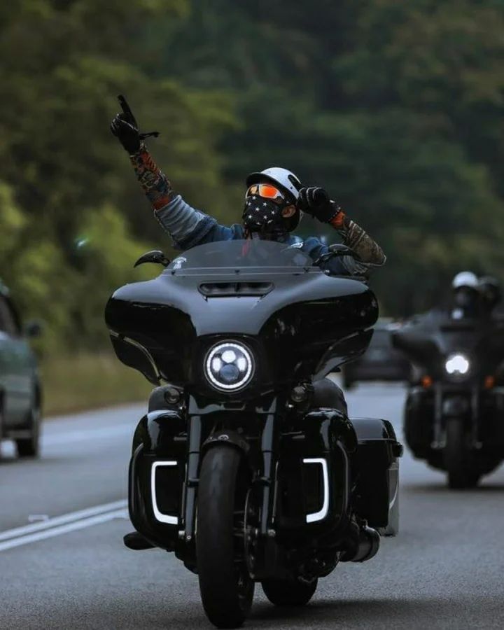 Motorcyclist with face covering rides a sleek black bike on a scenic road, confidently pointing forward.