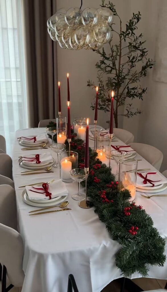 Elegant dining table set for Christmas with red candles, festive garland, and gold cutlery under a stylish glass chandelier.