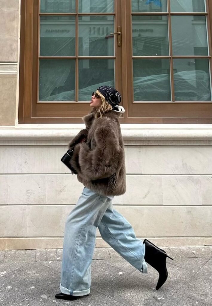 Fashionable woman in fur coat, wide jeans, and boots, walking stylishly by a window.