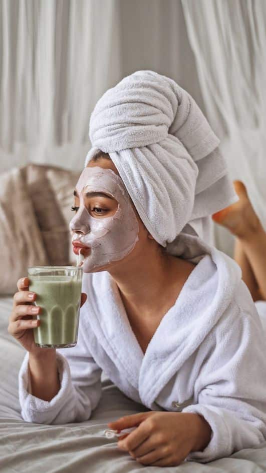 _ 8 Woman relaxing with a facial mask, towel turban, and drinking a green smoothie in a cozy bedroom setting. | Sky Rye Design Woman relaxing with a facial mask, towel turban, and drinking a green smoothie in a cozy bedroom setting.