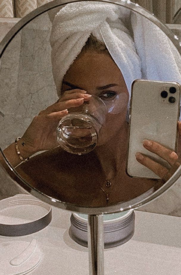 Woman in a towel head wrap drinks water, taking a mirror selfie with a smartphone, showcasing a relaxed skincare routine.