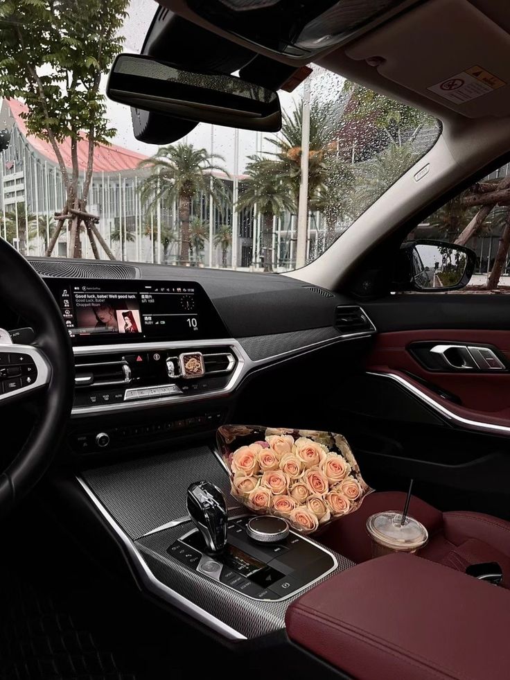 Luxurious car interior with red leather seats, touchscreen display, a bouquet of roses, and iced coffee. Raindrops on window.