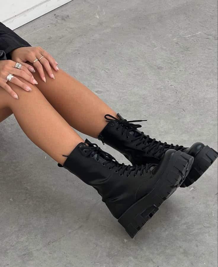 Woman wearing black combat boots, sitting on concrete floor. Stylish chunky footwear paired with manicured nails and silver rings.