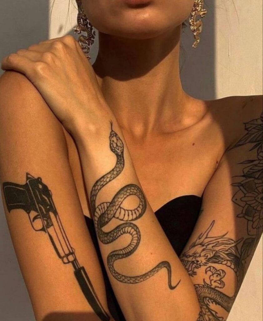 Woman with snake, gun, and dragon tattoos on her arms, wearing black top and intricate earrings, under warm sunlight.