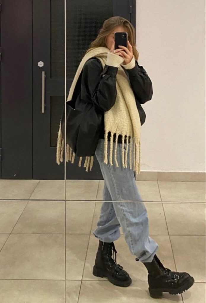 Fashion-forward individual in oversized scarf, black jacket, and boots taking a mirror selfie, showcasing a casual yet stylish look.
