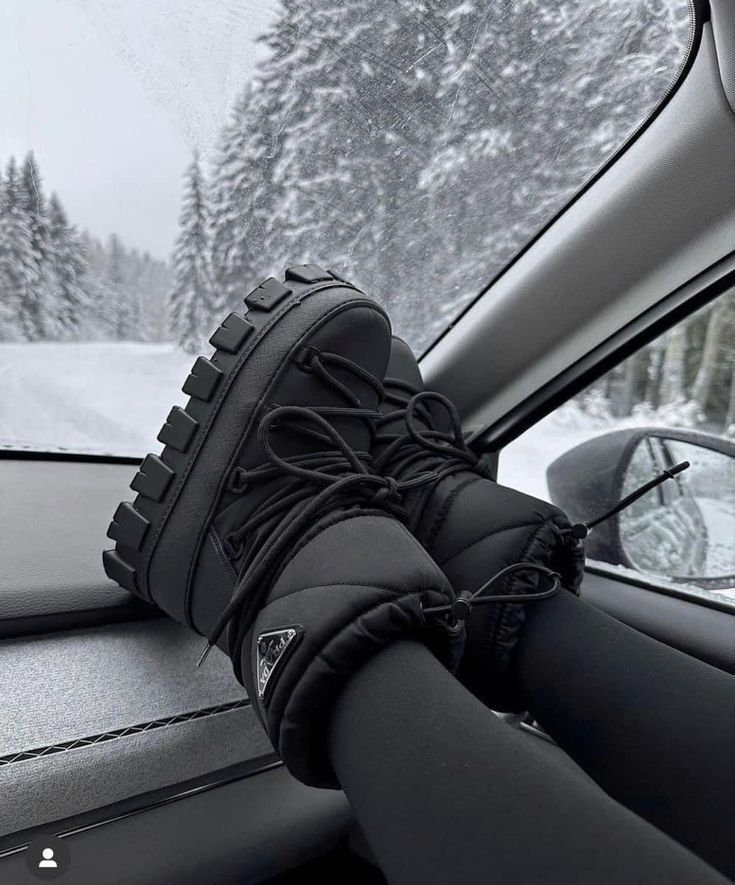 Black winter boots on car dashboard with snowy forest view in background, highlighting travel and winter fashion vibes.