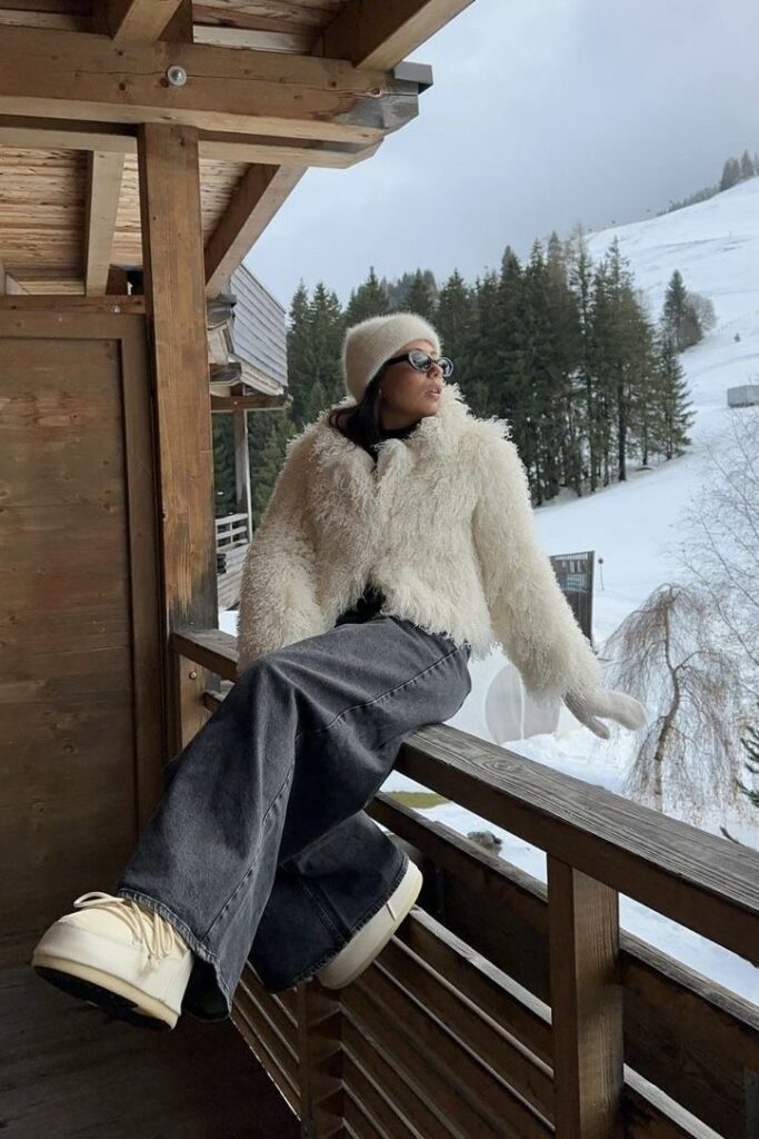 Woman in a fluffy coat and hat enjoys a snowy mountain view from a wooden balcony, dressed in winter fashion.