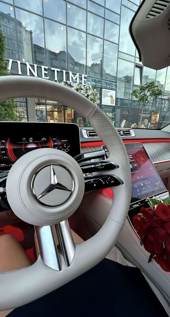 Luxury car interior with modern dashboard and steering wheel, parked outside a glass-fronted building. Red roses on the console.