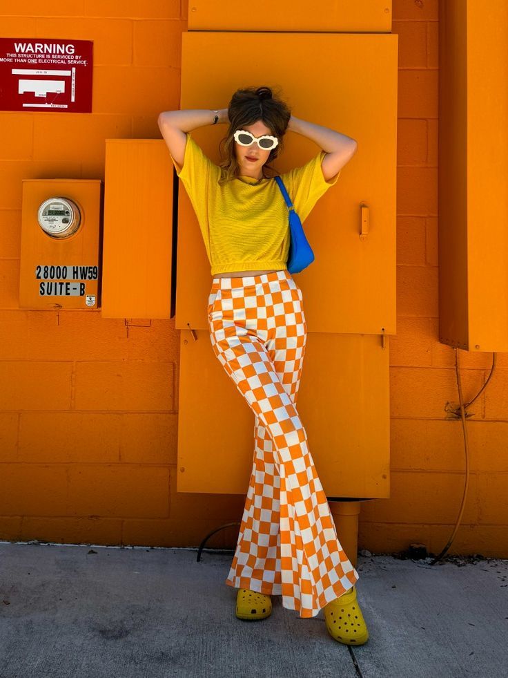 Stylish woman in yellow top and checkered pants poses against an orange wall, wearing sunglasses and holding a blue bag.