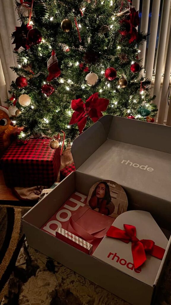 8d67bcfa-0776-4d74-94aa-14546814e1b3 Christmas tree lit with ornaments next to an open gift box labeled rhode, featuring red packaging and festive decorations. | Sky Rye Design Christmas tree lit with ornaments next to an open gift box labeled rhode, featuring red packaging and festive decorations.