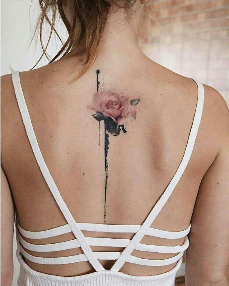 Back tattoo of a pink rose with abstract black ink lines on a woman wearing a white strappy top.