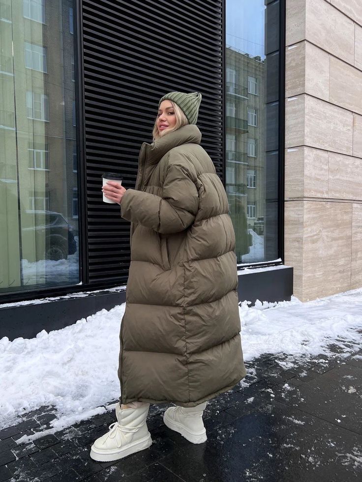 Woman in warm taupe puffer coat and beanie holding coffee on snowy city sidewalk.