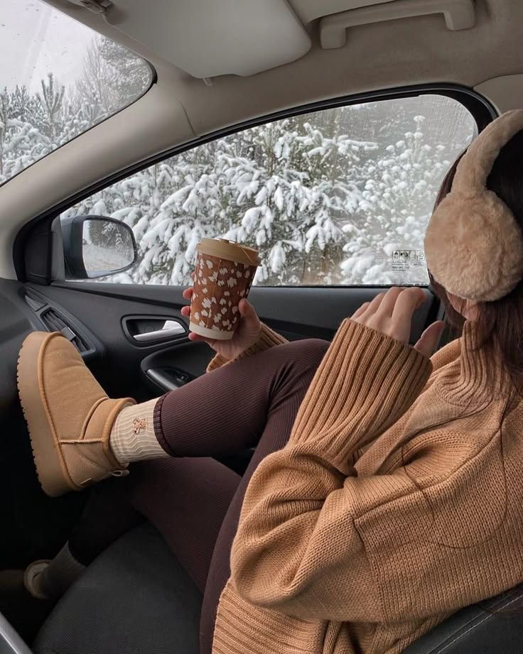 Cozy winter drive: person in car wearing earmuffs, sipping coffee, snowy forest view outside. Perfect for cold weather relaxation.