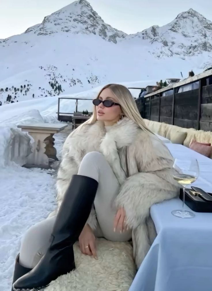 Fashionable woman in fur coat and sunglasses, relaxing with wine in a snowy mountain setting, showcasing winter style and elegance.