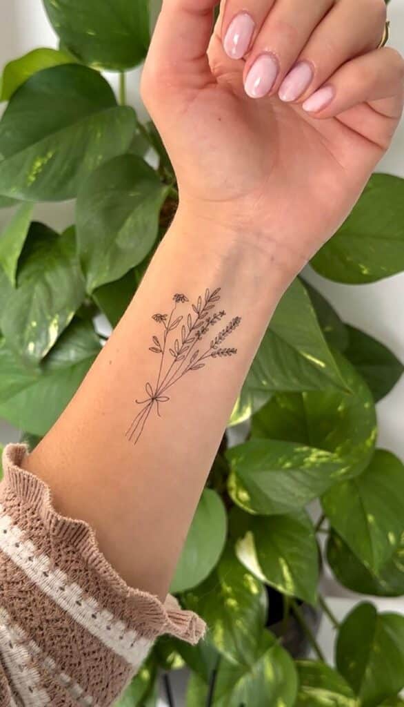 940d86f4-be62-48b3-8d5f-b29c3a429e16 Minimalist floral tattoo on a woman's forearm with pink nails, set against a background of vibrant green leaves. | Sky Rye Design Minimalist floral tattoo on a woman's forearm with pink nails, set against a background of vibrant green leaves.
