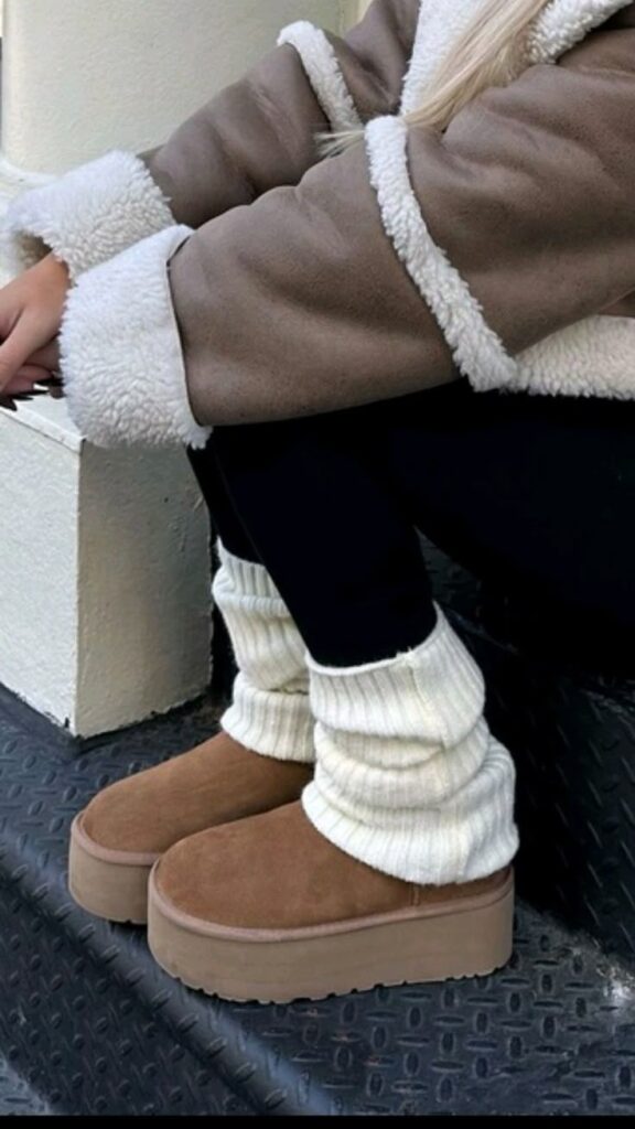 Cozy winter fashion: Woman in brown shearling coat, black leggings, and tan platform boots with white leg warmers on city steps.