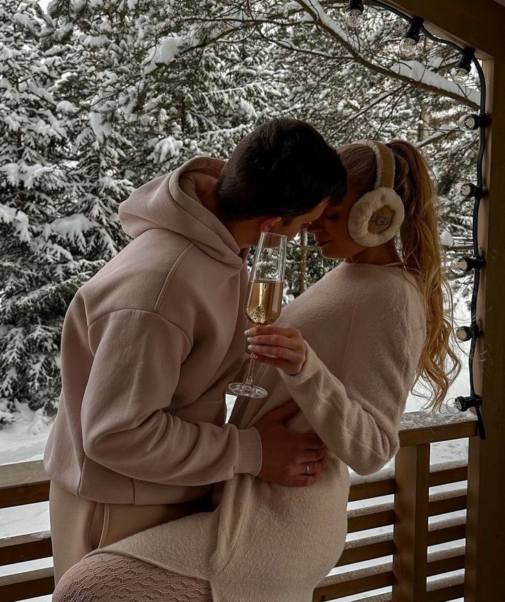 Couple in cozy winter attire shares a romantic moment on a snowy balcony, holding a glass of champagne, surrounded by a serene forest.