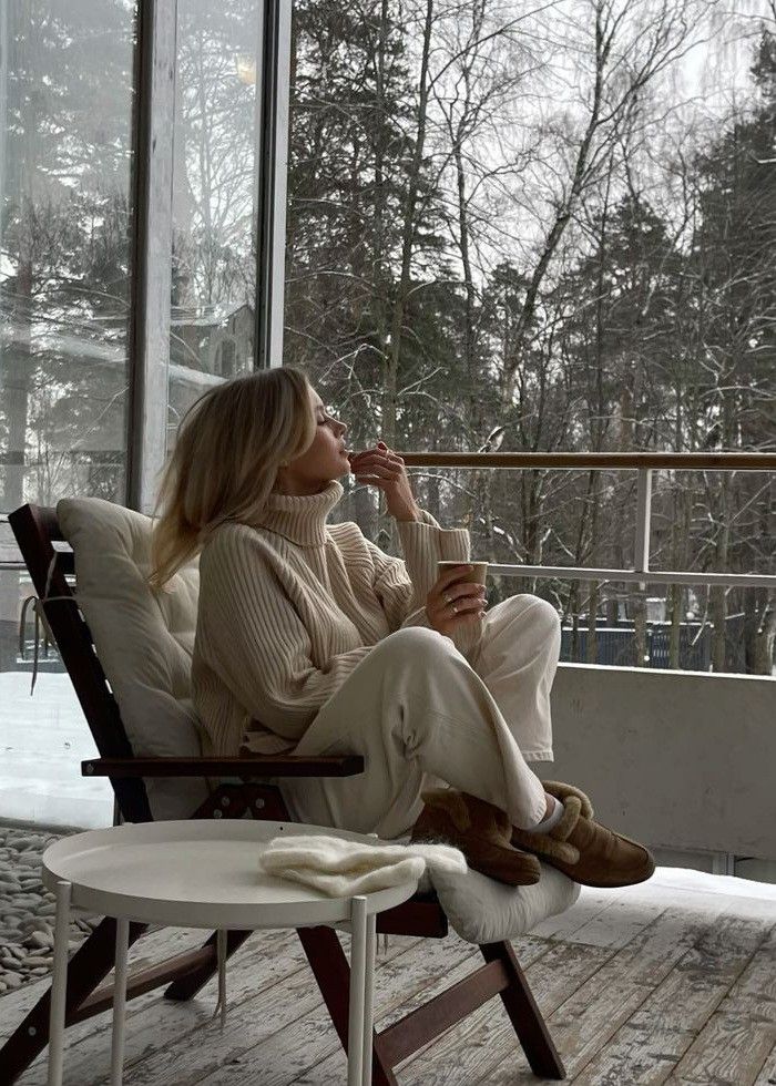 Cozy winter scene: Woman relaxing on a balcony in a sweater, sipping coffee, surrounded by snow-covered trees and a peaceful atmosphere.