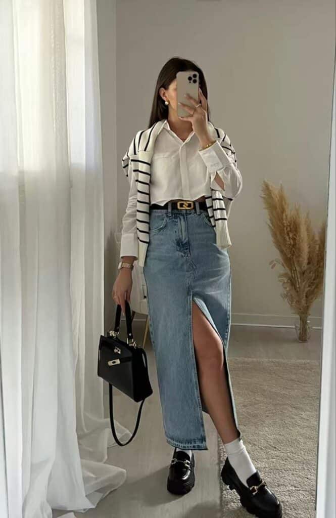 Stylish woman in white shirt, striped sweater, and denim skirt with a slit, holding a black handbag. Casual chic outfit inspiration.