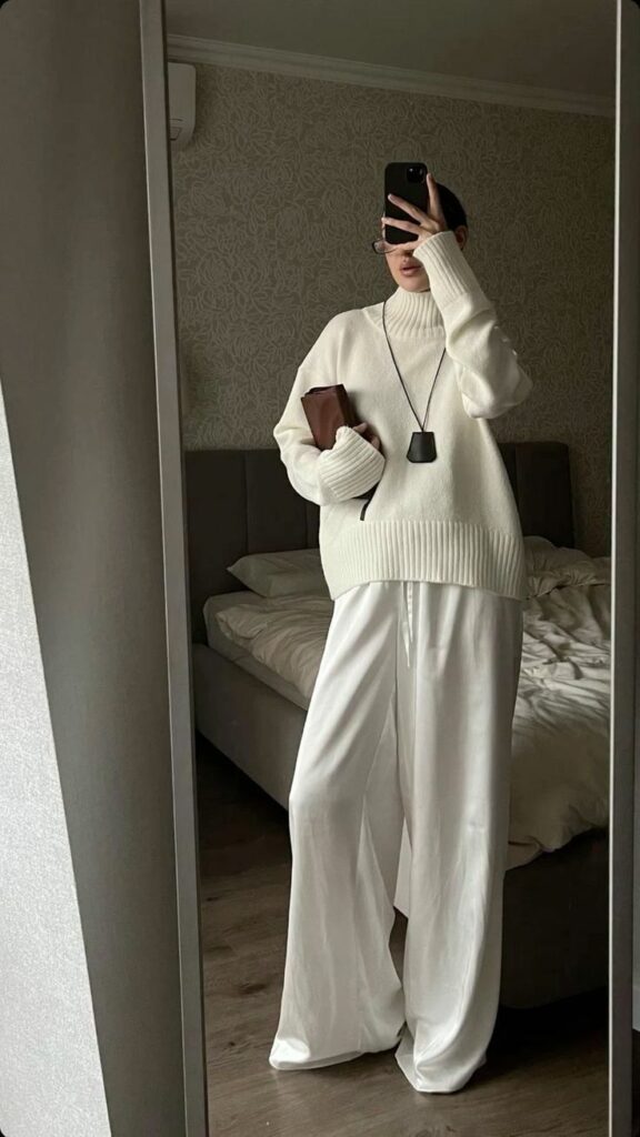 Person in cozy white turtleneck and wide pants, taking a mirror selfie, holding a clutch, in a minimalist bedroom setting.